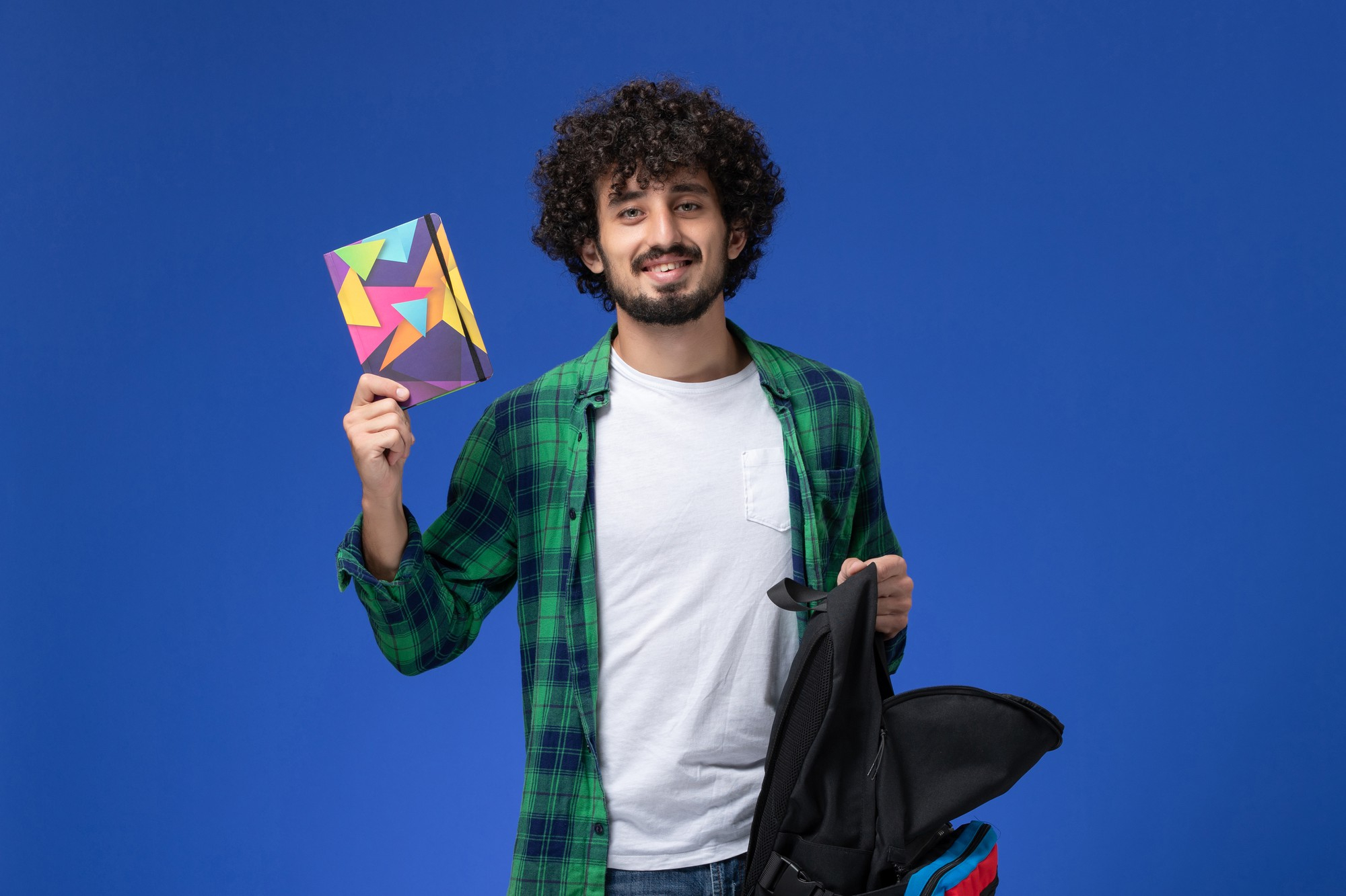 A student with a backpack holding up a notebook against a blue background